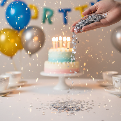 Silver metallic star confetti being poured from hand onto party table with birthday cake and balloons